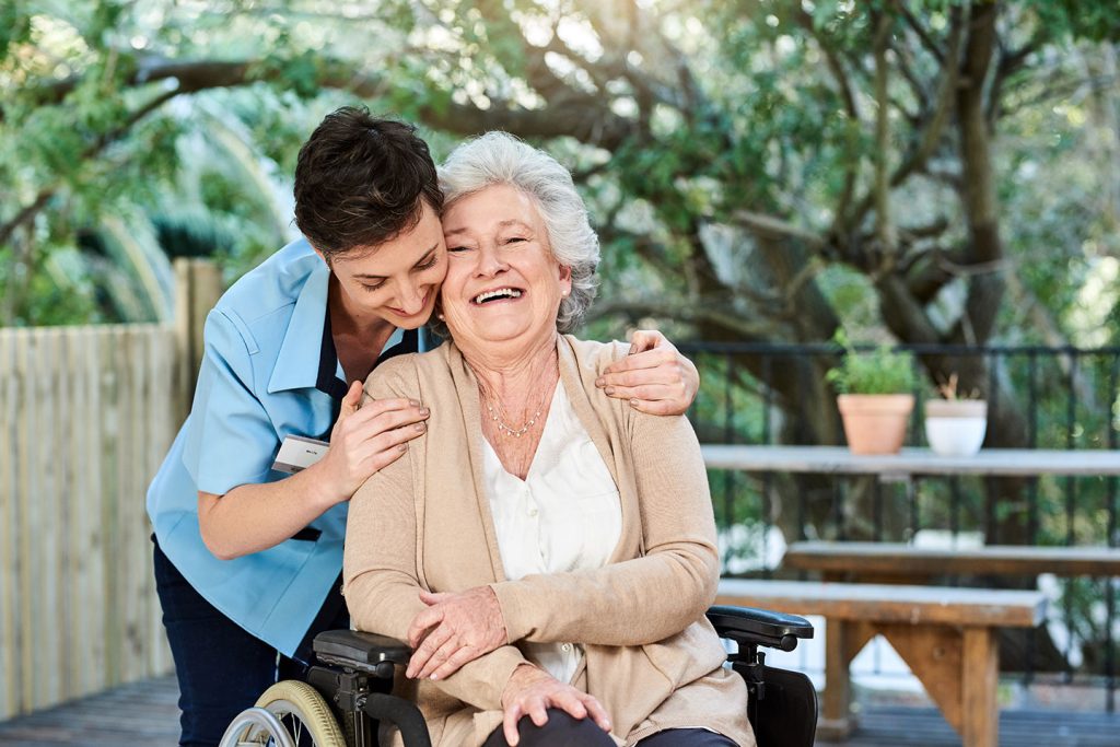A nurse in a blue uniform leaning down to hug a laughing older woman seated in a wheelchair on a garden deck