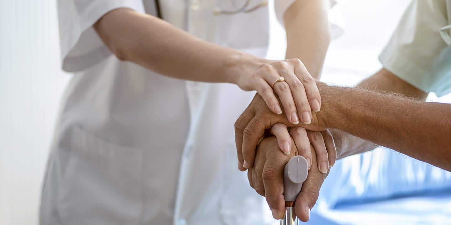 A nurse in scrubs rests a hand on an older person's hands, folded over the handle of a cane.