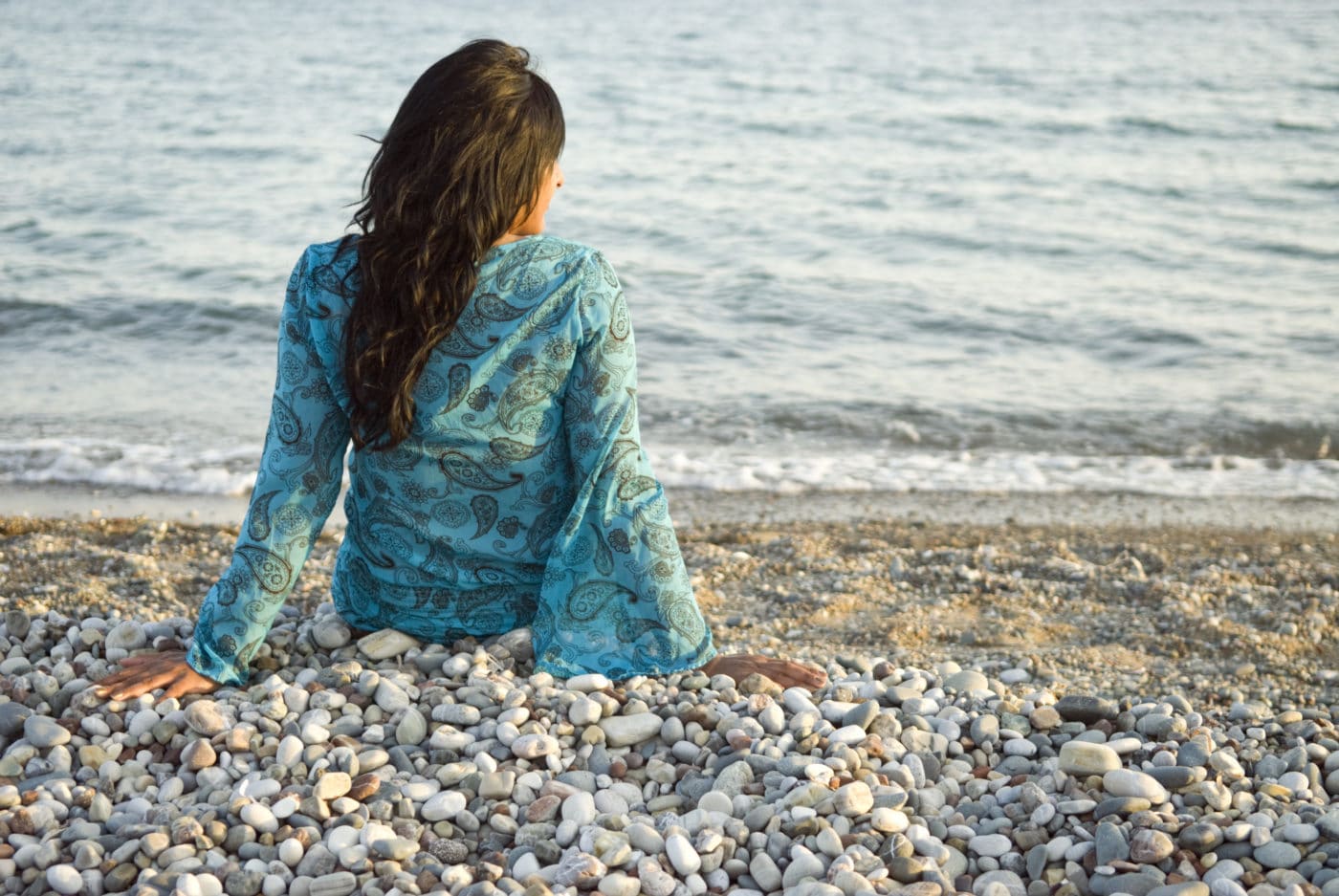 A woman sitting alone on a pebble beach, looking out at the sea