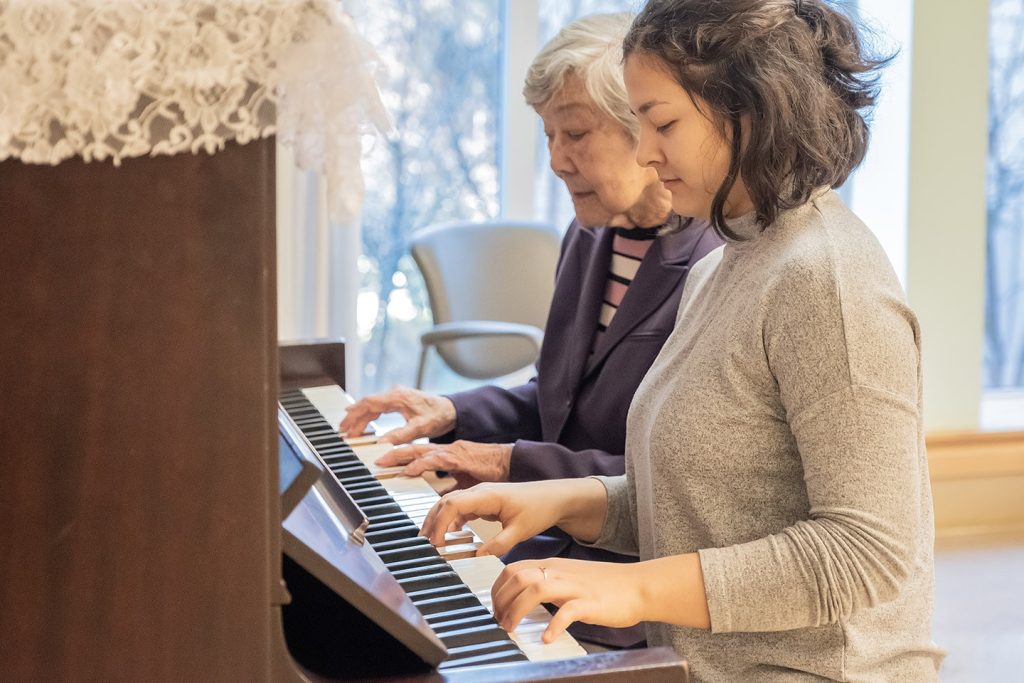 A music therapist and an older woman playing an upright piano together, their hands side by side on the keys
