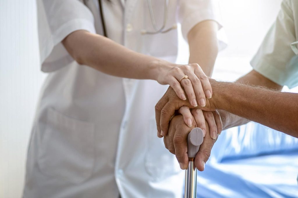 A doctor resting a hand over an older patient's hands, folded on the top of a walking cane