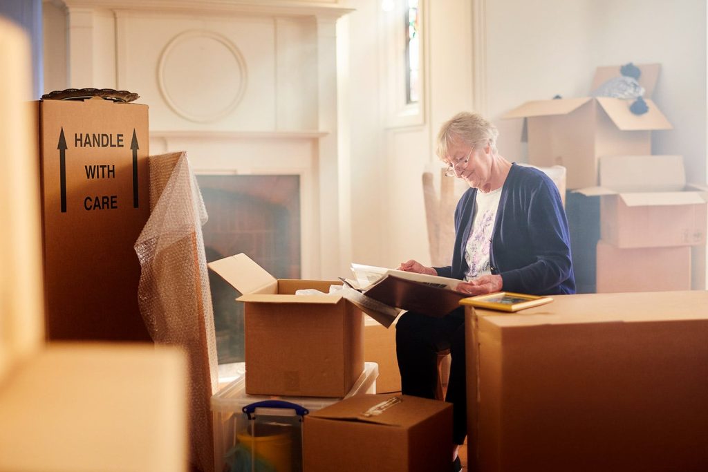 An older woman sorting framed pictures out of a packing box in a room stacked with cartons