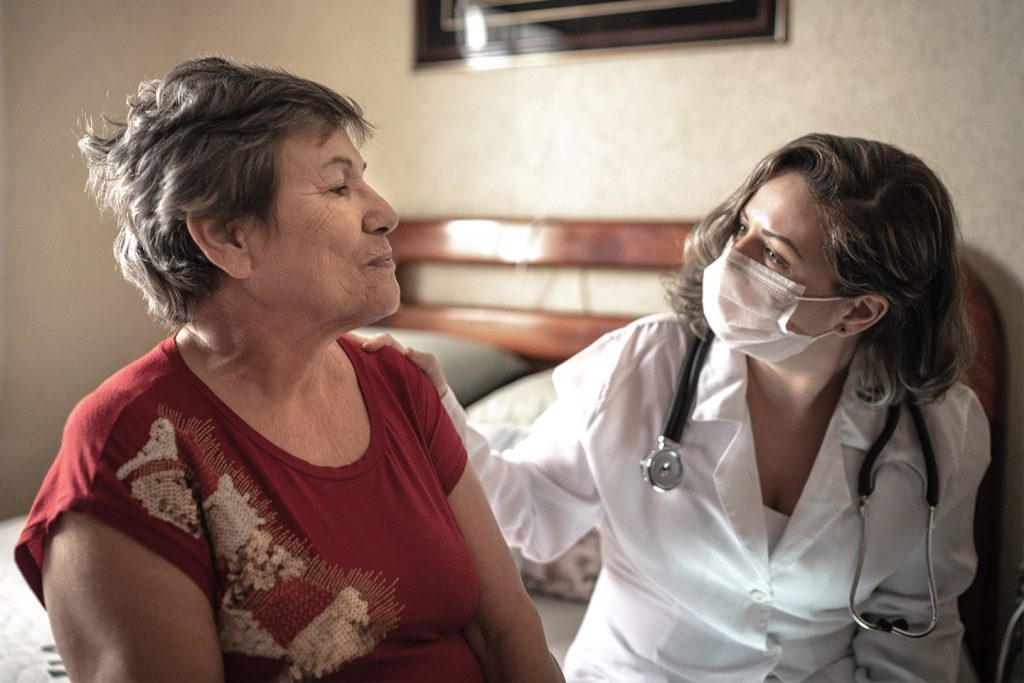 A clinician in a white coat and face mask sitting at a bedside, one hand on an older woman's shoulder