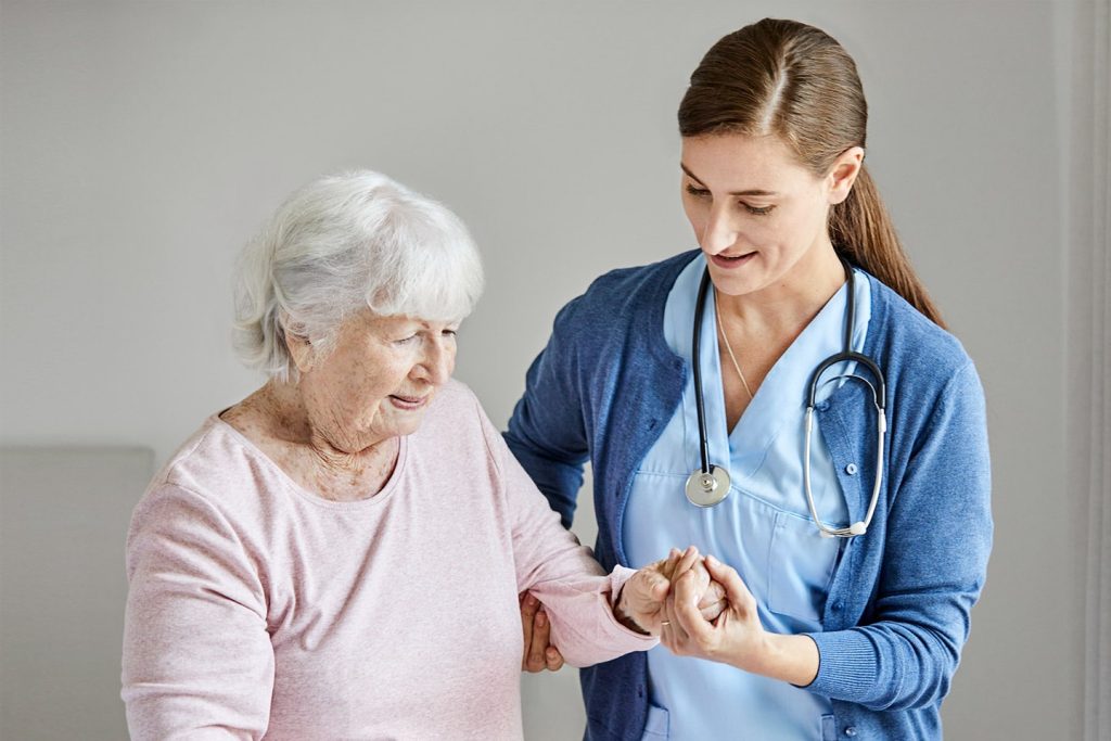 A nurse in scrubs steadying an older woman by the hand as they walk