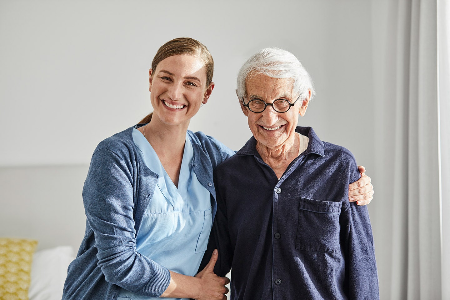 A nurse in blue scrubs with her arm around a smiling older man in his room