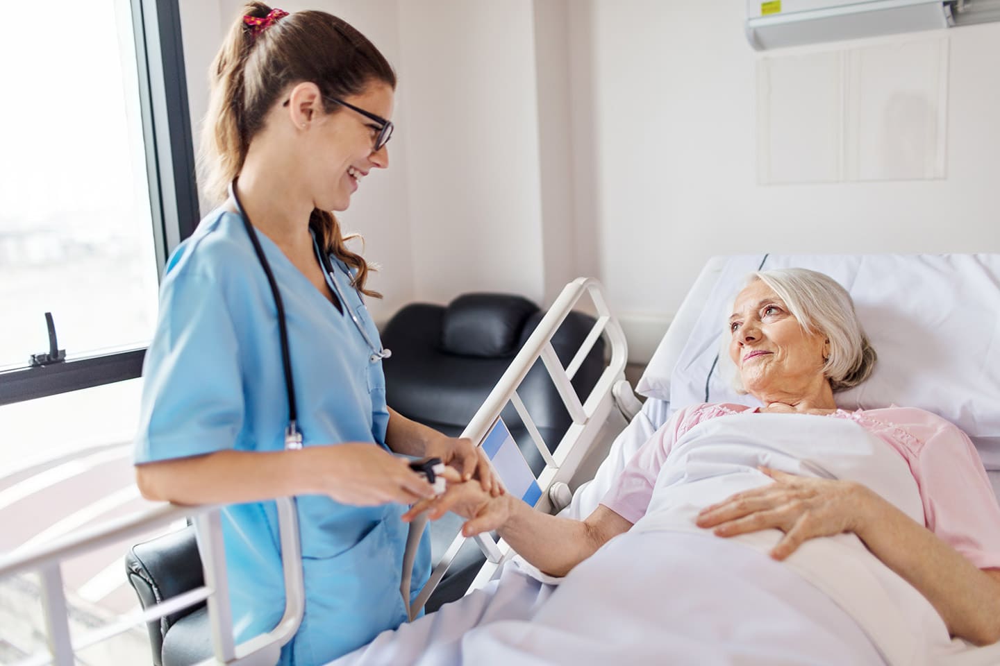 A nurse in scrubs holding the hand of a smiling patient resting in bed
