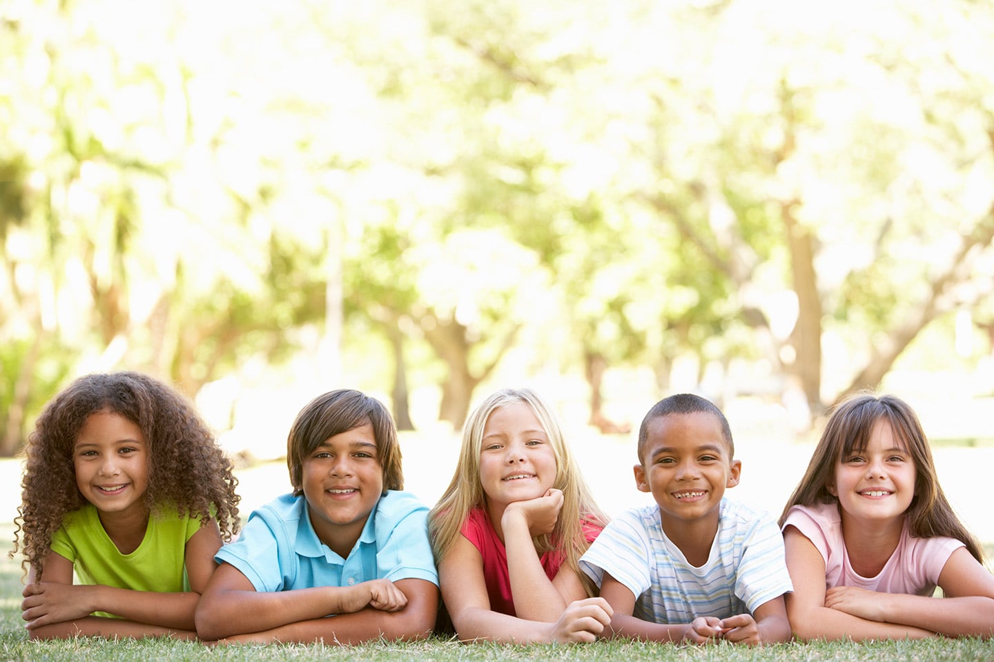 Five children lying side by side on the grass in a park, propped on their elbows and smiling