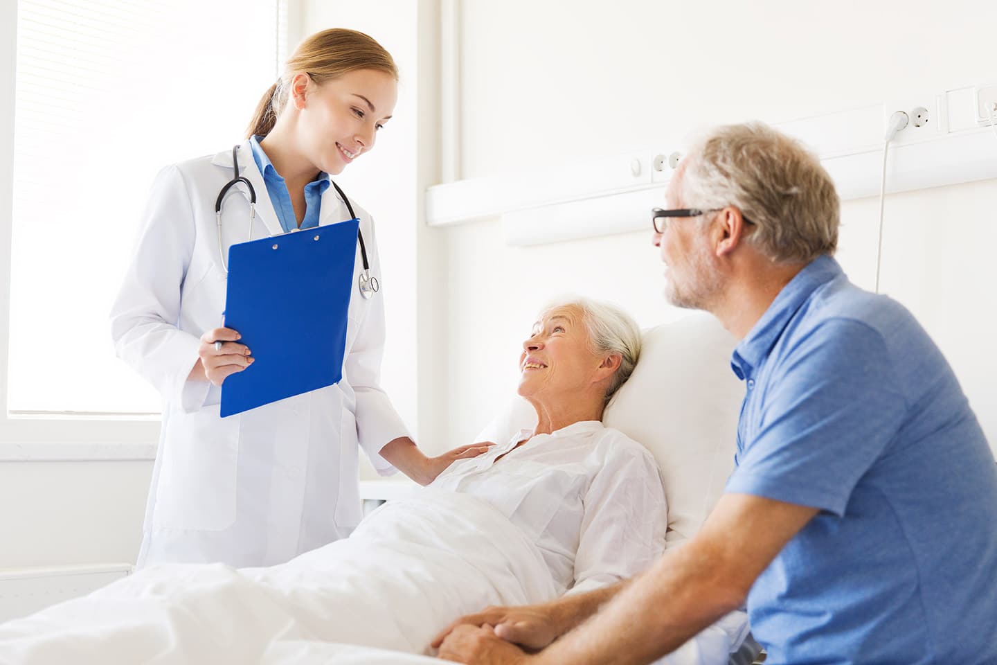 A nurse with a clipboard at the bedside of a smiling older woman, a man in a blue polo shirt sitting beside her