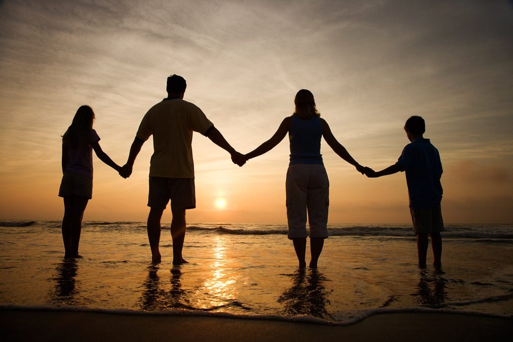 A family of four holding hands at the water's edge, silhouetted against the setting sun