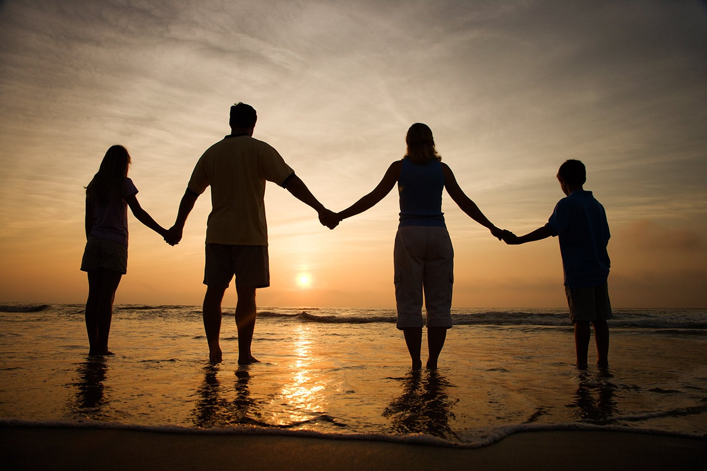 A family of four holding hands at the water's edge, silhouetted against the setting sun
