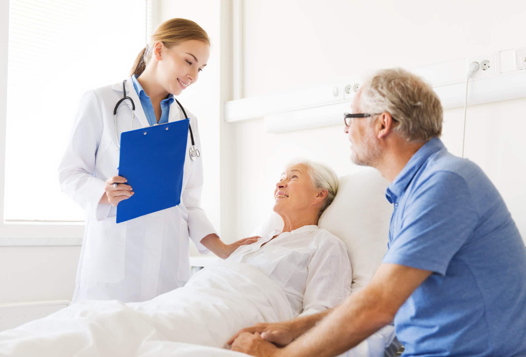 A visitor holds a patient's hand at her bedside while a clinician talks with them both