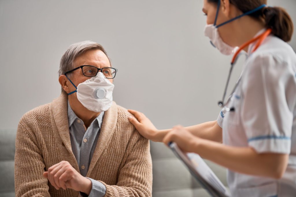 An older man in a face mask sitting with a nurse, who rests a hand on his shoulder