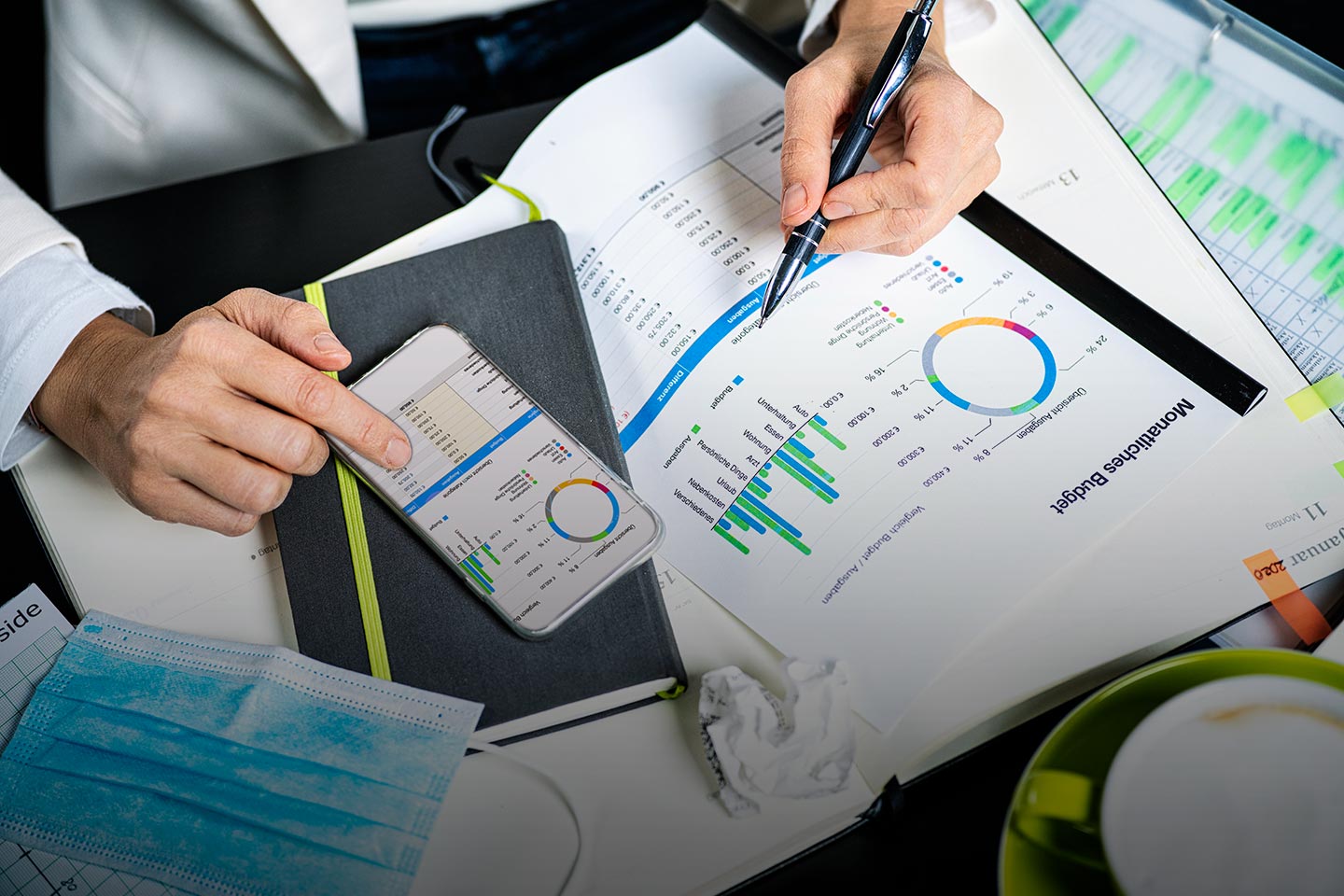 Hands annotating a printed monthly budget chart beside a phone showing the same figures.
