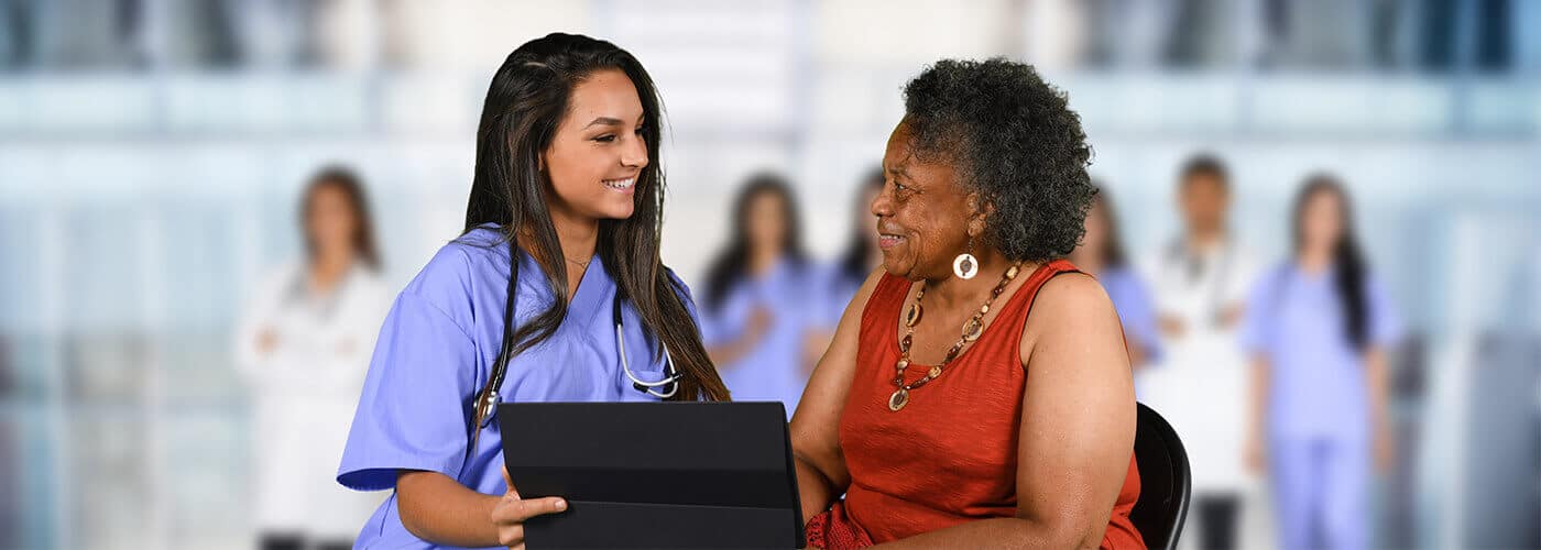 A nurse in scrubs holding a chart smiles with an older woman seated beside her, colleagues standing behind them.
