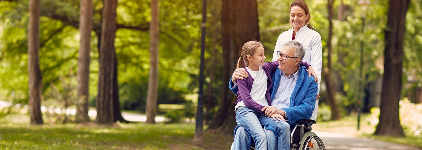 A nurse walking an older man in a wheelchair through a park while his granddaughter sits on his lap with an arm around him