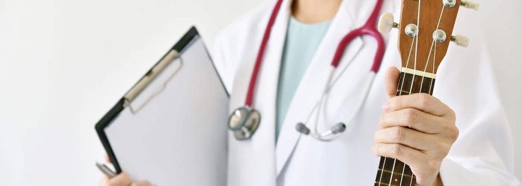 A clinician in a white coat and stethoscope holding a clipboard in one hand and the neck of a ukulele in the other