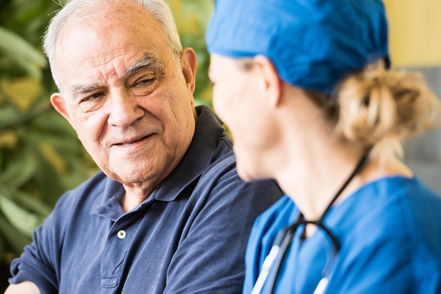 A nurse in scrubs with a stethoscope in conversation with an older man, listening as he speaks