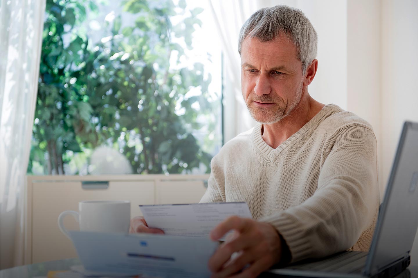 A man at a kitchen table reads a printed statement beside an open laptop.