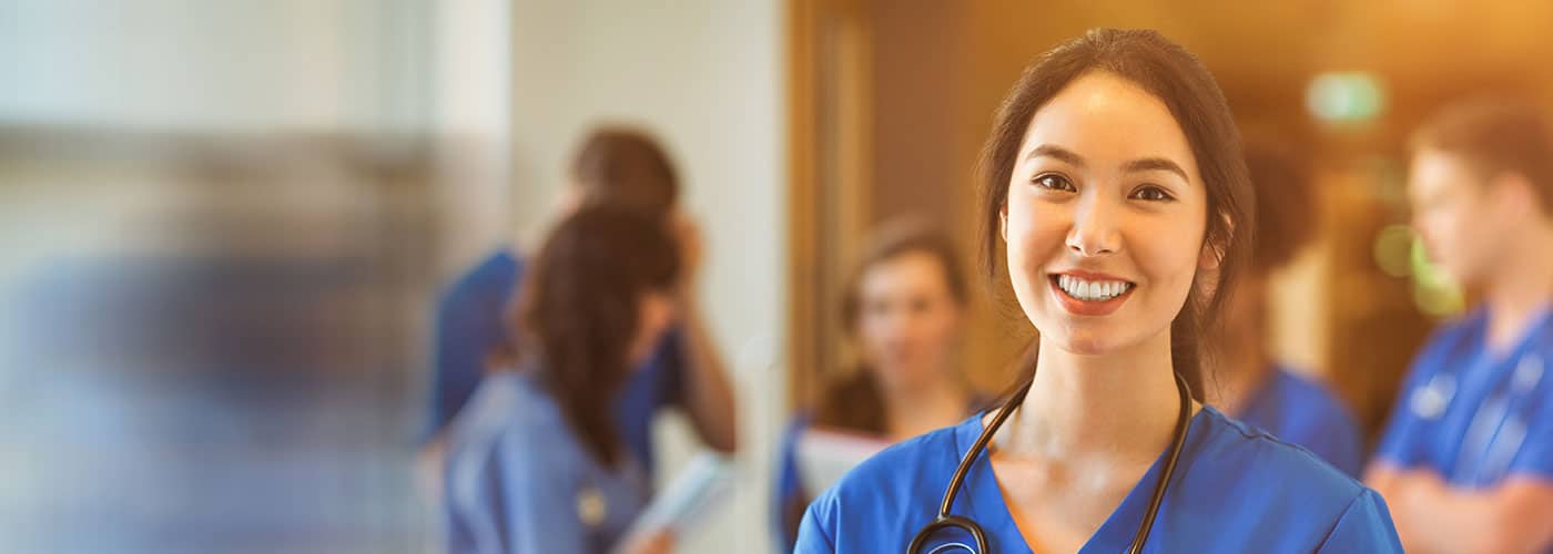 A smiling clinician in blue scrubs with a stethoscope, colleagues talking in the corridor behind her