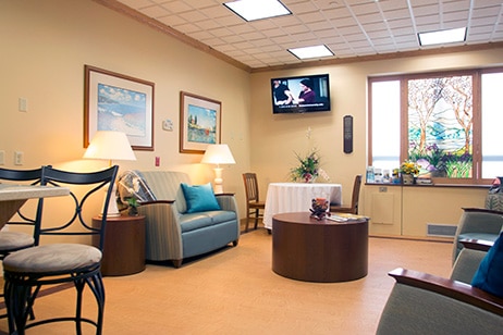 The family room at the Bethesda Memorial Care Center, with armchairs, table lamps, framed seascapes and a wall-mounted television