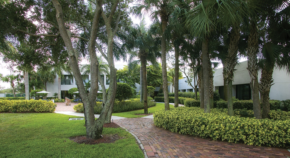 A brick walkway curving through a landscaped garden between palm trees and clipped hedges, beside a care center building with shaded seating.