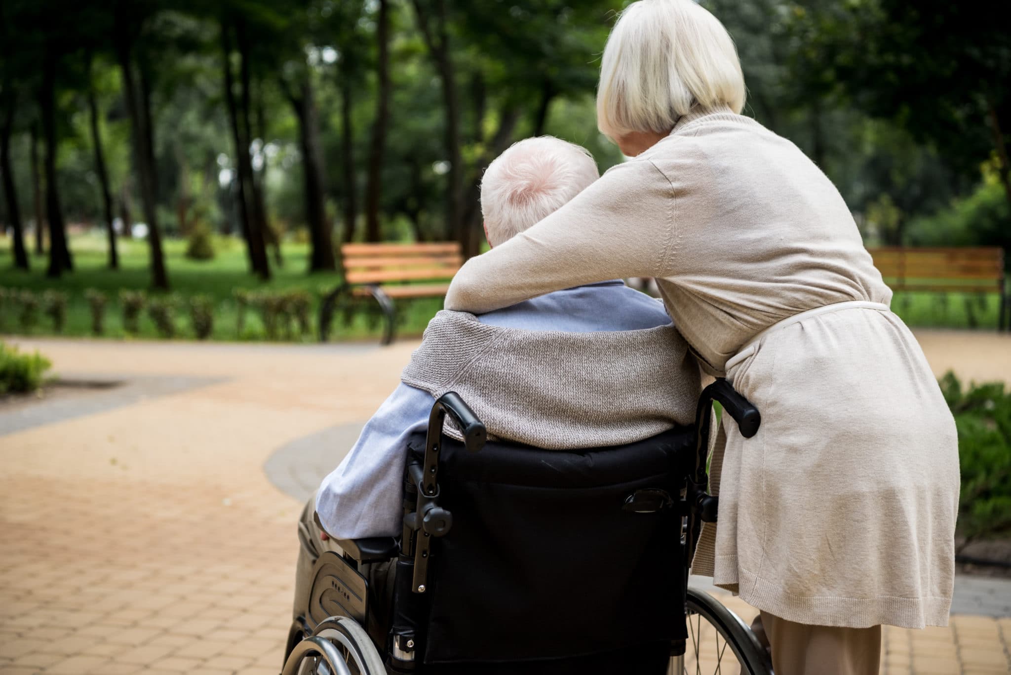 An older woman leaning over to embrace a man seated in a wheelchair on a tree-lined park path
