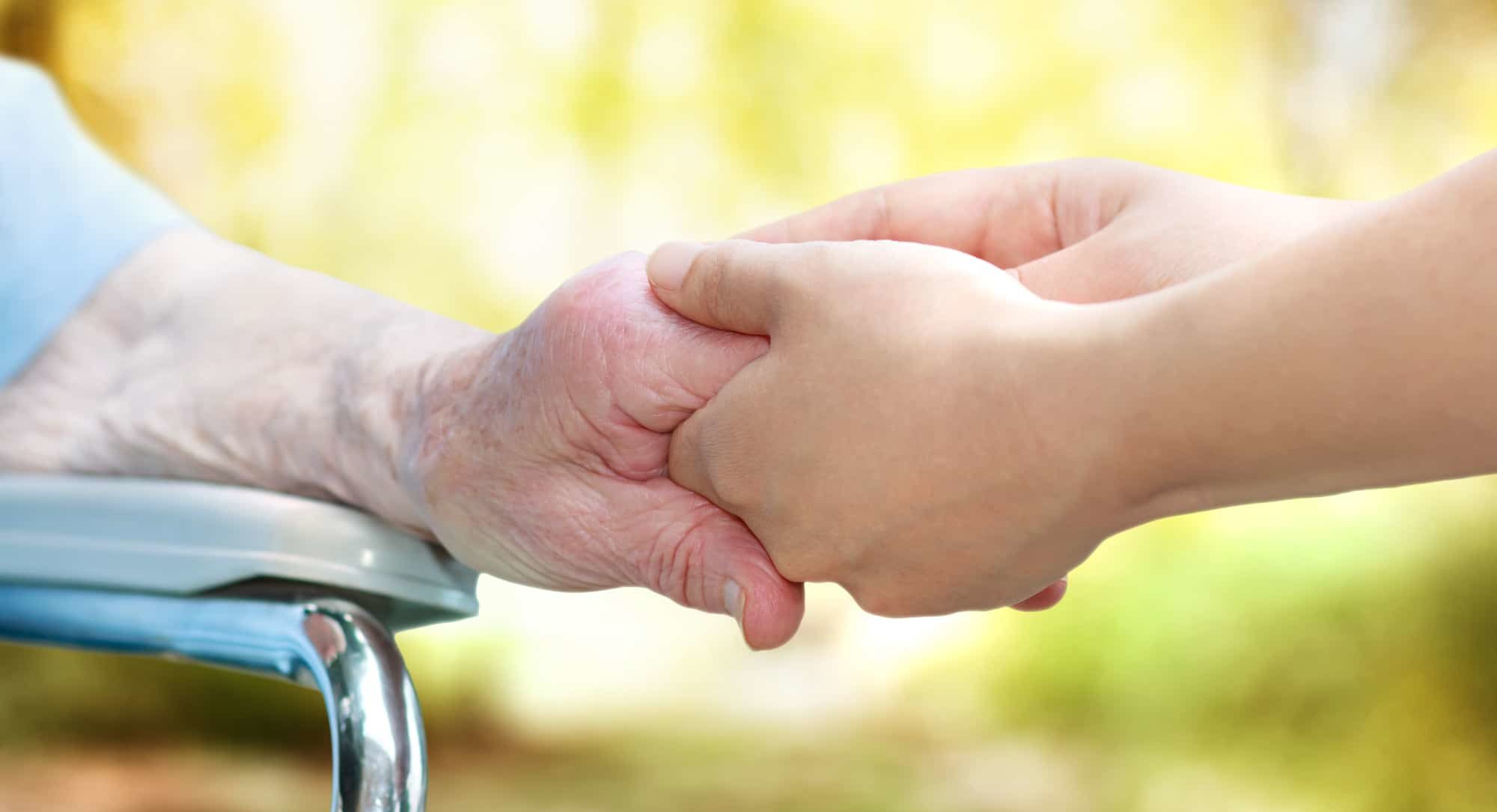 A younger hand holding the hand of an older person seated in a wheelchair