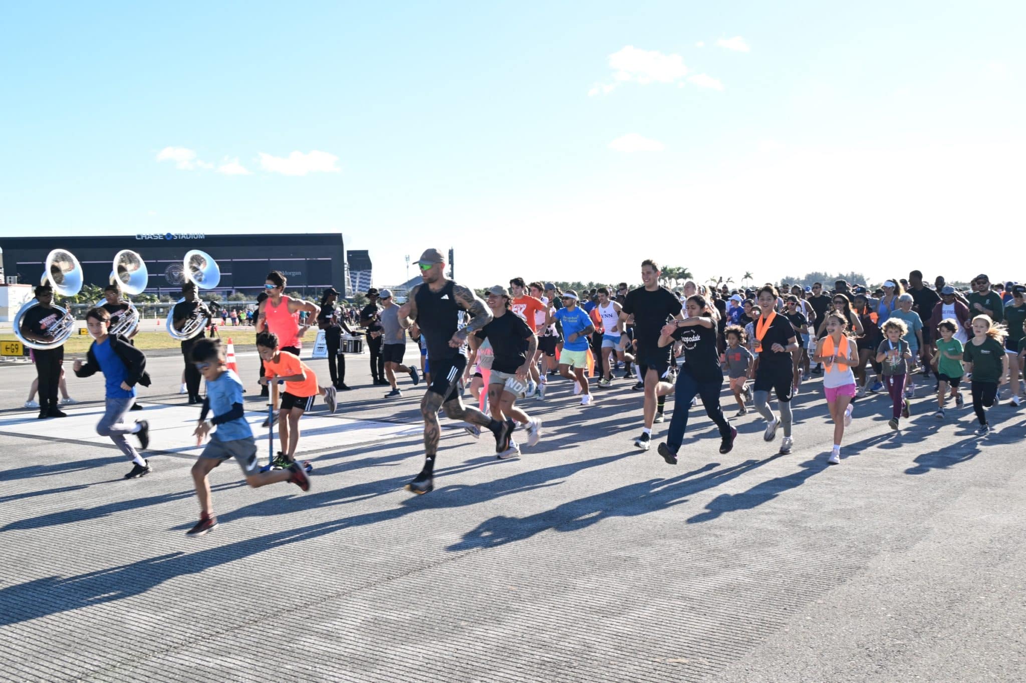Runners setting off along the airport runway at the start of the Uptown 5K on the Runway, with a marching band playing beside the start line