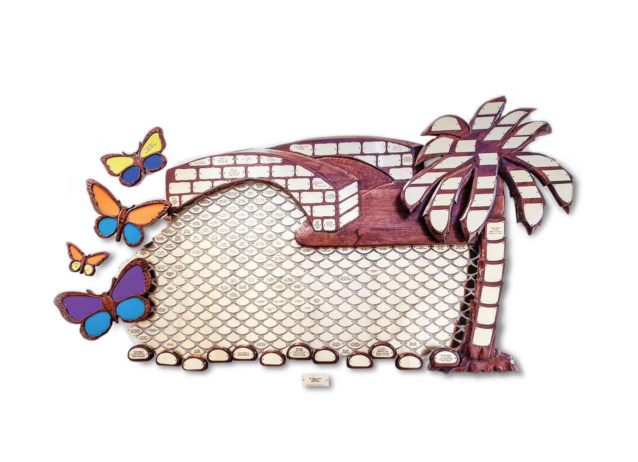The Butterfly Bridge Memorial: a carved wooden wall sculpture of an arched bridge and a palm tree, edged with stained-glass-style butterflies and covered in small engraved name plaques.