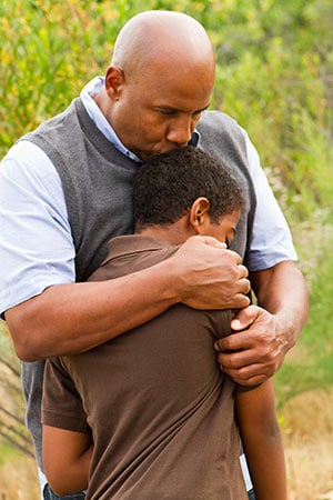 A father holds his young son close, his head resting against the boy's, outdoors.