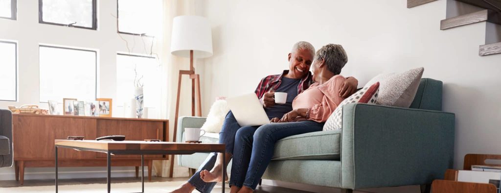 An older couple sitting close together on a sofa at home, sharing a laptop and a cup of coffee