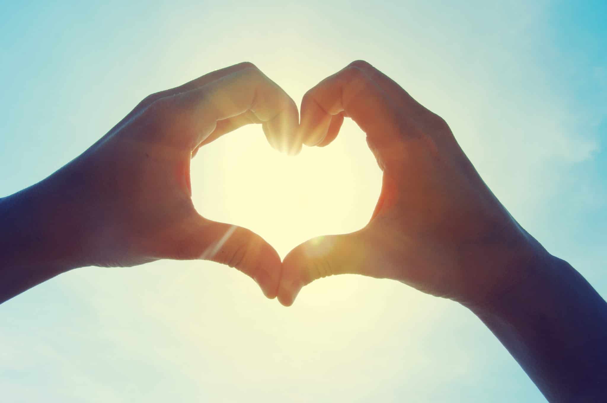 Two hands framing the sun in the shape of a heart against a bright sky