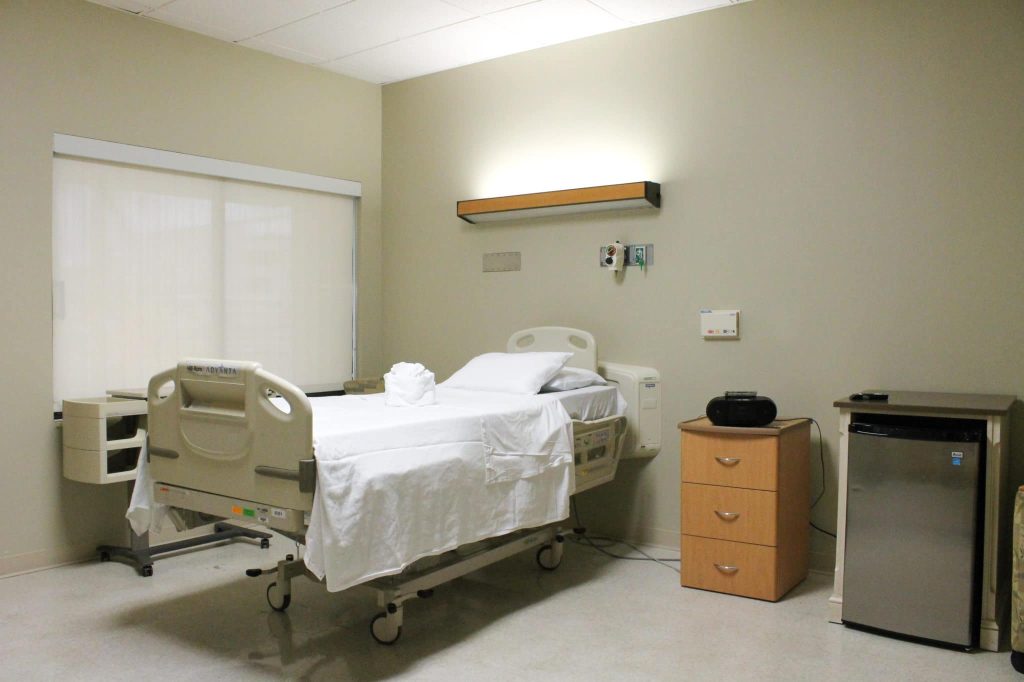 A patient room at the Delray Medical Center care center, with a made hospital bed, a bedside cabinet and a shaded window