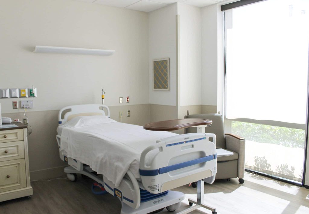 A patient room at the Jupiter Care Center, with a hospital bed, an overbed table, a recliner and a full-height window onto greenery