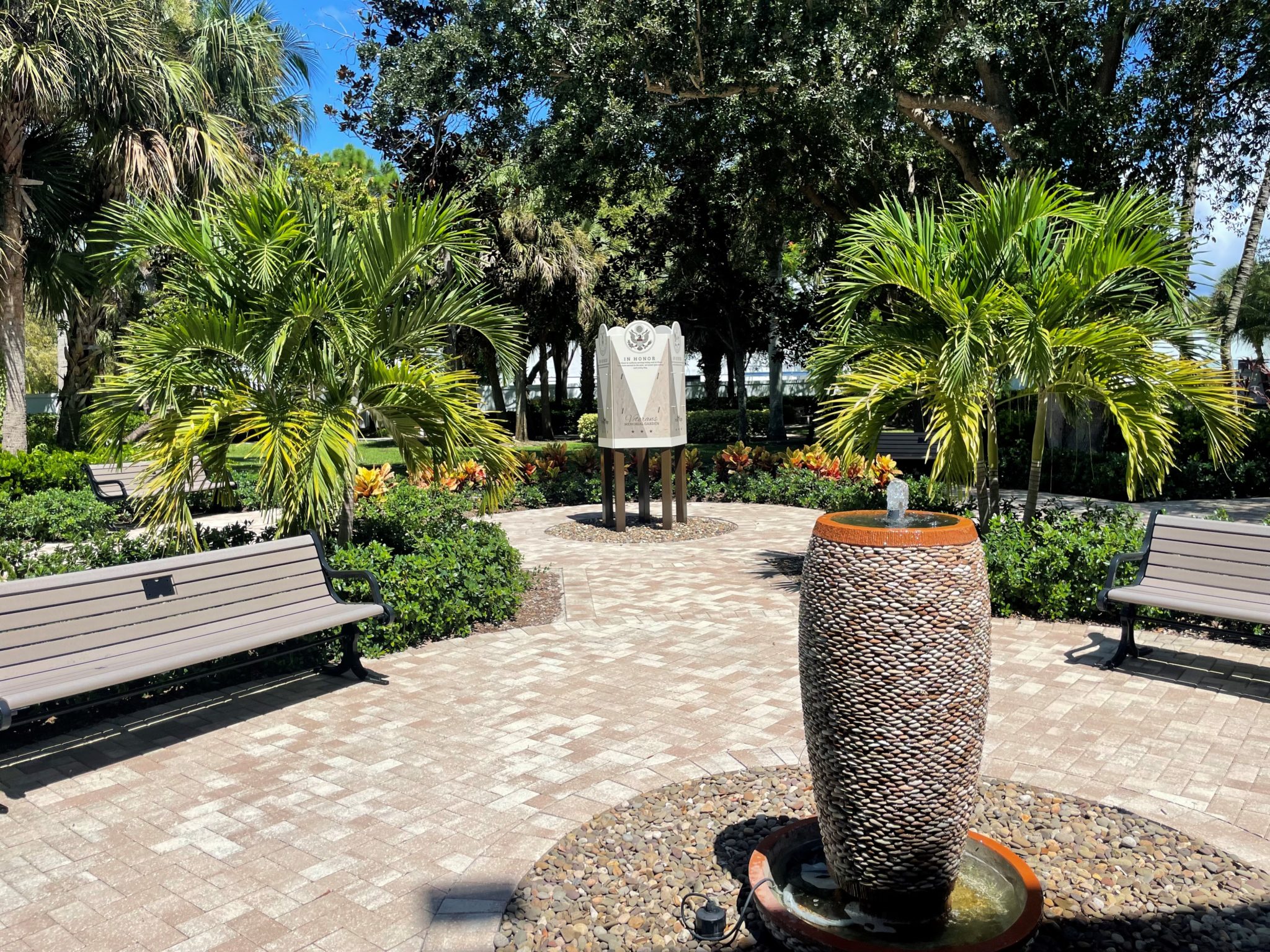 A paved garden circle with benches, palms and a pebble-mosaic fountain, with a memorial marker set among the plantings