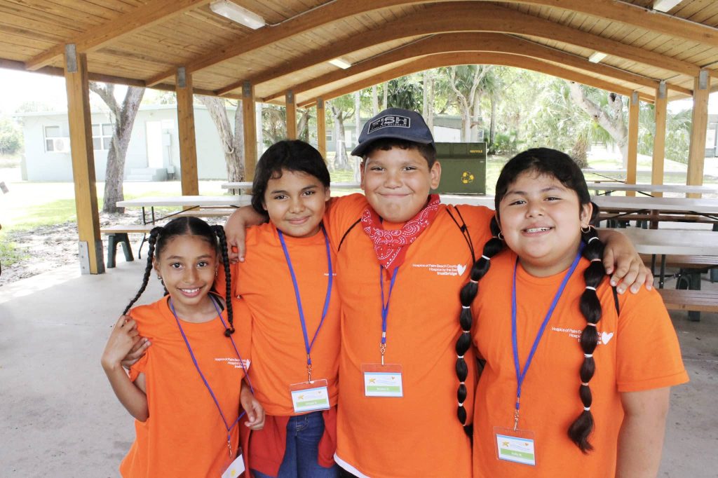 Four children in orange Camp Good Grief T-shirts standing arm in arm under a picnic pavilion