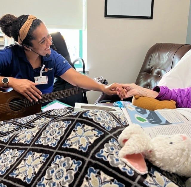 A Trustbridge music therapist with a guitar holding an older patient's hand at her bedside