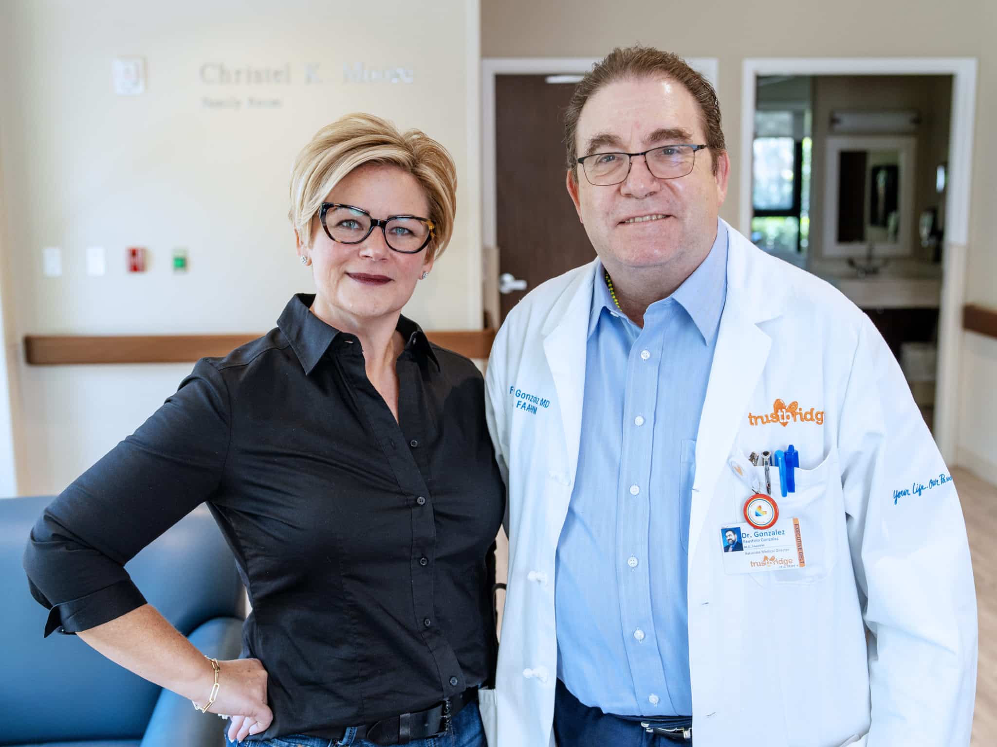 Heather and Dr. G standing together in a Trustbridge family room; he wears a white coat with the Trustbridge logo.