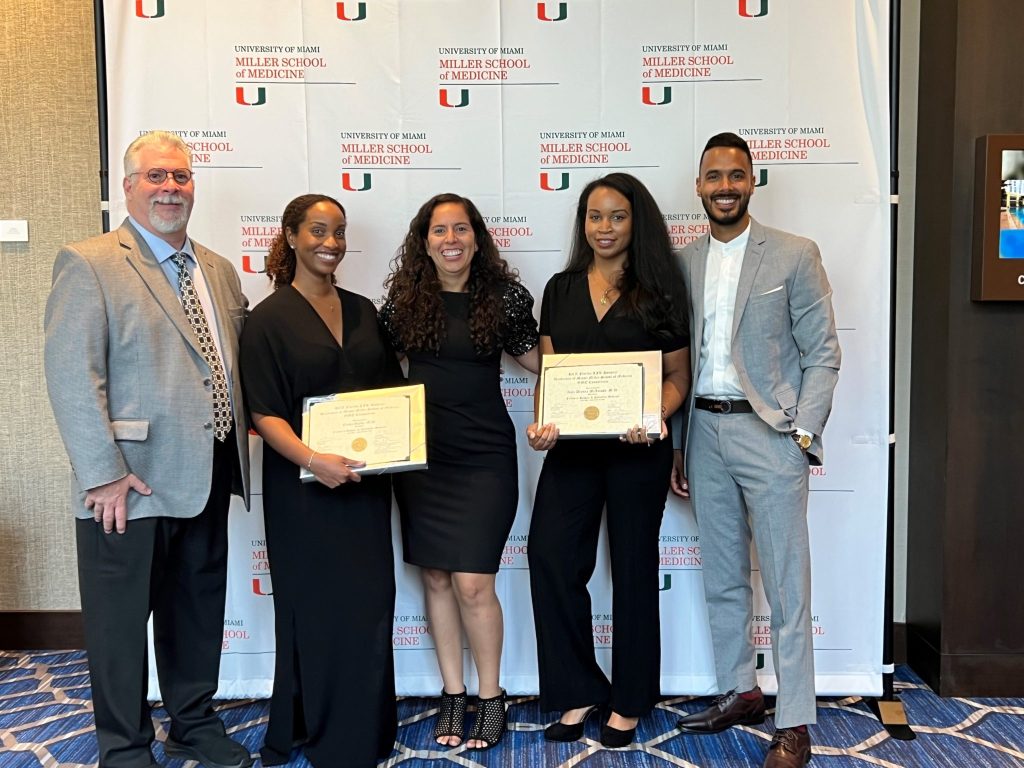 Two graduating fellows holding their certificates with three colleagues, in front of a University of Miami Miller School of Medicine backdrop