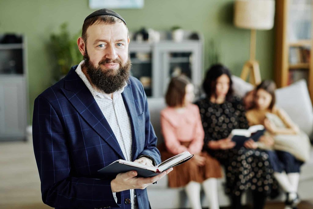 A rabbi in a kippah holding an open book, with a mother and two children reading together on a sofa behind him