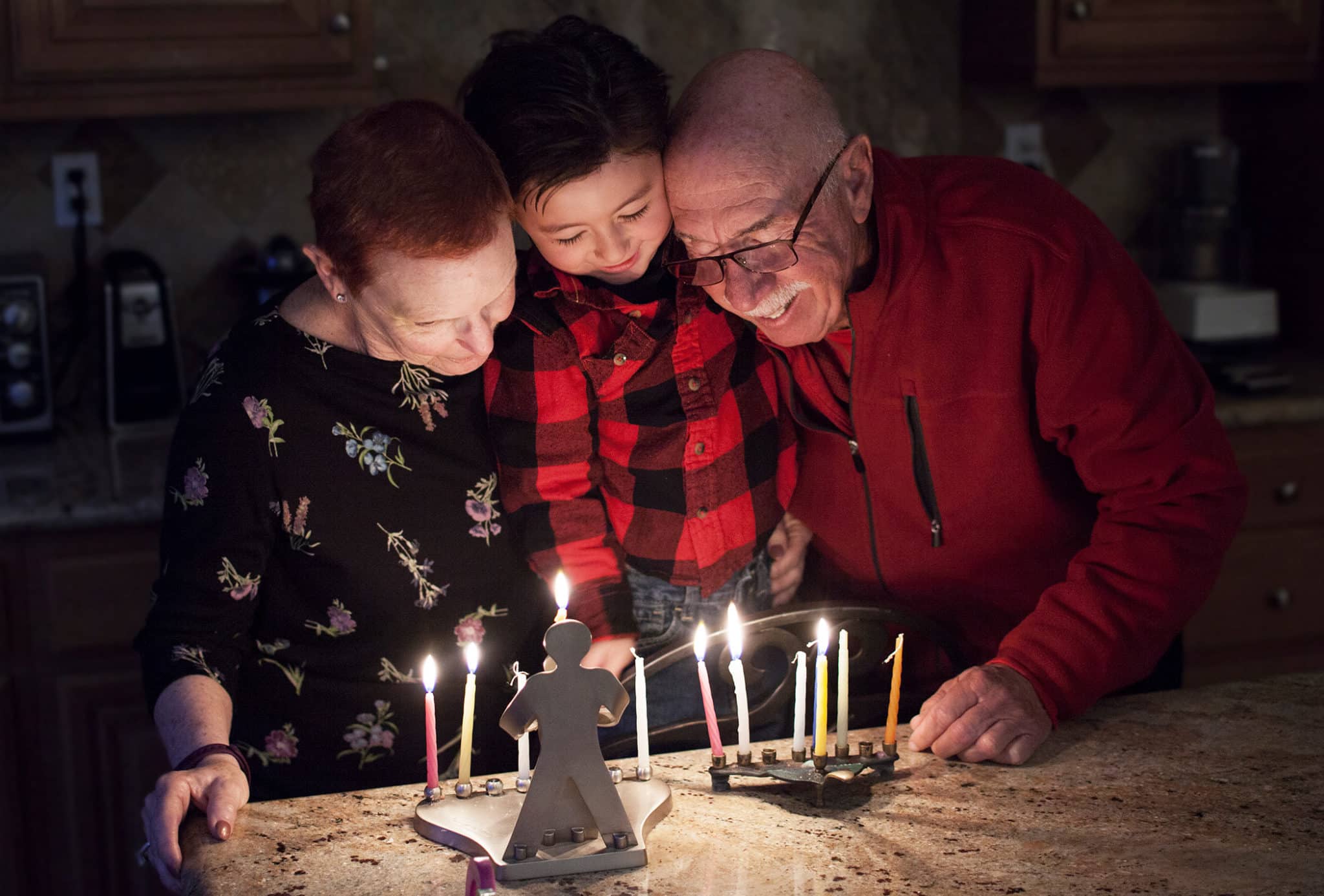 A grandmother, grandfather and young boy leaning close together as they watch two lit menorahs on a kitchen counter